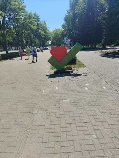 This media file captures a sunlit scene in Salihorsk, Belarus. The foreground is dominated by a paved walkway, with a large red heart sculpture featuring green lettering visible on the right. The lettering appears to spell out "СОЛИГОРСК", the name of the city.
In the background, several people are walking along the path. On the left, two women are walking side-by-side, one in a pink top and red pants, the other in a green top and beige shorts. Behind them, a man in a blue shirt and shorts walks away from the camera, while another man in a white shirt walks further in the distance. Lush green trees line the walkway on the left and right, creating a pleasant park-like atmosphere. The sky is a clear, bright blue, suggesting a warm, sunny day. The overall mood is relaxed and leisurely, characteristic of a pleasant day in a public outdoor space.