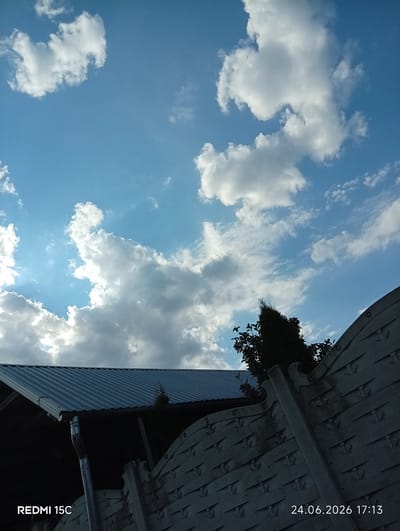 This is a photograph taken outdoors, likely in a residential or semi-rural area. The scene is dominated by a bright blue sky filled with scattered white and gray clouds. Sunlight is visible peeking through some of the clouds, suggesting it is either early morning or late afternoon.
In the foreground, a gray corrugated metal roof of a building is visible, with a downspout attached. To the right of the roof, a textured gray fence with a repeating decorative pattern runs diagonally across the frame. Behind the fence, the dark silhouette of a tall, narrow evergreen tree is visible, along with some other foliage. The overall setting appears to be a yard or garden area.
The time of day is indicated by the timestamp "24.06.2026 17:13" in the lower right corner of the image, suggesting it was taken in the late afternoon. The weather appears to be partly cloudy, with the sun still shining. There is no discernible activity or event taking place; the image is a still shot of the environment.
The text "REDMI 15C" is visible in the lower left corner, likely indicating the model of the phone used to take the photograph. The location context of Mykolaivka, Ukraine, places the scene geographically. No people are visible in the image, and the overall mood is tranquil and serene.