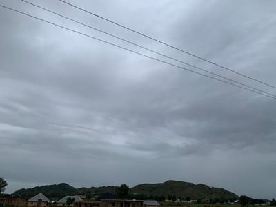 An overcast sky looms over a landscape of rolling, tree-covered hills dotted with buildings. Three dark power lines stretch diagonally across the upper portion of the frame. In the foreground, several structures are visible, some appearing to be under construction with exposed brickwork and incomplete roofs. Two houses with grey, pitched roofs stand out amongst the other buildings. The overall impression is a rural or semi-urban setting in Zawan, Nigeria, under a cloudy sky. No people are discernible in the image.