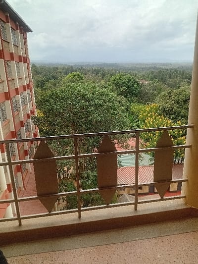 The image captures an elevated exterior view from a balcony in Karingani ward, Kenya, under an overcast sky. The foreground features a light-brown, speckled terrazzo floor. A light-colored metal railing, composed of horizontal bars and vertical supports, spans the width of the frame, featuring decorative, shield-shaped inserts with pointed top and bottom edges. A cream-colored structural pillar is visible on the right edge. To the left, a multi-story building with a facade characterized by alternating horizontal bands of reddish-brown and cream-colored sections, punctuated by numerous windows, extends upward. Beyond the railing and immediately below, scattered rooftops with green and reddish-brown corrugated iron are partially visible amidst a dense canopy of various green trees. The midground and distant background are dominated by an extensive, undulating landscape of dark green foliage, indicative of heavy vegetation, fading into a hazy horizon line under a uniformly gray, cloudy sky. No human subjects, animals, or discernible actions are present.