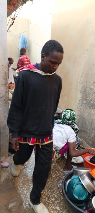 The image captures a candid moment in a narrow outdoor corridor or alleyway, likely within a residential compound in Kaura Goje, Nigeria. The walls are light-colored and appear to be made of concrete or mud brick, with a corrugated metal roof visible in the upper left corner.
In the foreground, a young Black man walks towards the left side of the frame. He is dressed in a black hooded sweatshirt with a red and white lining visible in the hood, worn over a blue collared shirt. A distinctive band of red, blue, and yellow striped fabric is visible beneath the hem of his sweatshirt, possibly an underlying garment. He wears black pants and light-colored, slip-on shoes or clogs. His head is slightly bowed, and his expression is neutral.
To the right, partially obscured by the foreground man, a woman is seen from behind, squatting or kneeling. She is engaged in washing dishes, immersing her hands in an orange basin. She wears a white t-shirt, a vibrantly patterned skirt or wrapper in green, pink, and yellow, and a matching green and white patterned head covering. Various metal and plastic bowls are visible in a larger black basin next to her.
Further in the background, to the left, another man in a red and white striped shirt stands near a young child. The child is wrapped in a white cloth or towel and stands beside a brown woven basket. A light blue piece of cloth hangs on a line strung across the alleyway. The ground is wet and appears to be a mix of concrete and pebbles. The bright lighting suggests it is daytime. There is no visible text in the image. The scene depicts routine domestic activities in a bustling outdoor setting.