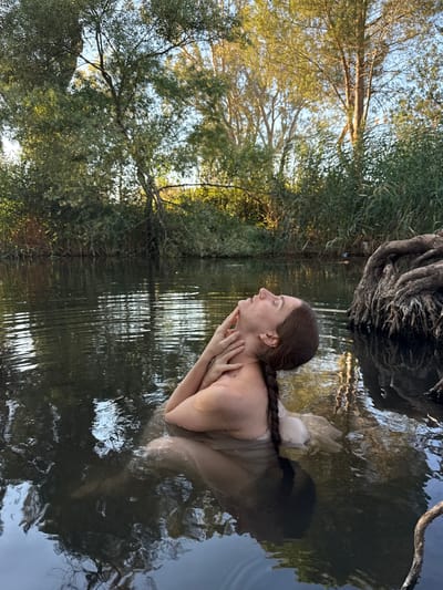 A woman with a braid is submerged in dark water, with her head tilted back and eyes closed, her hands gently touching her neck and jaw. She is wearing a light-colored, flowing garment that is also submerged. The water is calm with ripples reflecting the surrounding trees and sky. On the right side of the image, the exposed roots of a large tree emerge from the water. The background consists of dense green foliage and tall trees, illuminated by soft, warm light. The scene appears to be a natural water body, possibly a river or lake, surrounded by lush vegetation. Given the context of Katzrin, Israel, this setting is consistent with the natural landscapes found in the Golan Heights region, which includes wooded areas and water sources.
