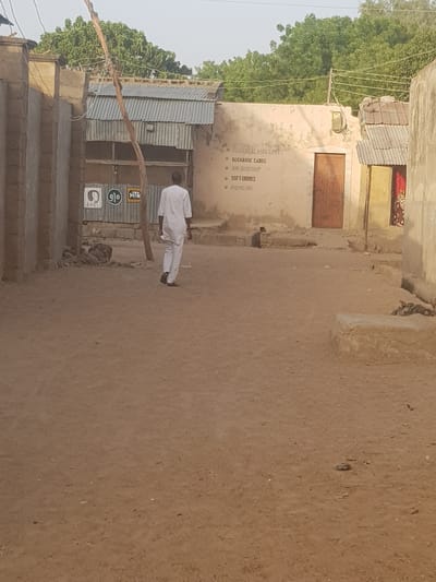 The scene is a dusty street in Dutsinma B, Nigeria. The environment appears arid with vegetation to the left. Buildings on both sides of the street have walls of earthen material with corrugated metal roofs. A man is walking along the street toward the right, wearing light-colored clothing. On the left, a building has signs for "Recharge Cards", "Soft Drinks", and "Provisions" painted on its exterior. Telecommunication signs are present on a building adjacent to it. The ground is a compacted dirt surface.