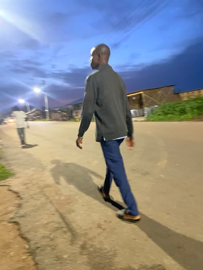 The image depicts an outdoor scene at dusk in Barikin Jirga Summa, Nigeria. A man is walking away from the camera on a paved road. He is wearing a dark grey long-sleeved shirt over a light blue collared shirt, and dark blue trousers. His right hand is visible, with a gold-colored bracelet on his wrist. He is wearing dark-colored open-toed sandals. A second person, wearing a white shirt and blue jeans, is walking in the opposite direction in the background, further down the road. The road is bordered by sparse vegetation on the left and buildings on the right. Streetlights are visible in the distance, illuminating the scene and casting long shadows. The sky is a deep blue with streaks of clouds. The overall atmosphere suggests a quiet evening in a semi-urban or rural setting.