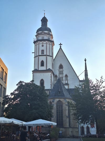The image depicts the historic Nikolaikirche (St. Nicholas Church) in Leipzig, Germany, bathed in the soft light of what appears to be late afternoon or early evening. The towering Gothic church, with its distinctive white facade and ornate steeple, dominates the center of the frame. A large, dark green tree partially obscures the lower portion of the church. In the foreground, a row of white outdoor umbrellas, bearing the text "Brauerei • Restaurant • Pizzeria," shades a busy outdoor seating area. Several people are visible sitting at tables and walking around, engaged in dining or socializing. The sky is a clear, pale blue, indicating fair weather. To the left of the church, a modern building with large windows is visible, contrasting with the historical architecture.