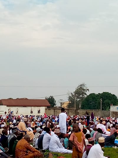 The image shows a large gathering of people in an outdoor area in Beni, Democratic Republic of Congo (coordinates: 0.489, 29.463). The people are sitting on the ground, mostly facing towards the right side of the image, likely participating in a religious gathering. Many of the men are wearing white clothing, and some are wearing hats. There is a low concrete wall in the background with a brick building behind it. Trees and a cloudy sky can be seen in the background.  There is an information board visible to the right with a sign that reads "PLAISTY MAISTER". The people are facing towards the right, but the viewer's perspective is facing them from behind.  The grassy area is sparsely covered with vegetation.
