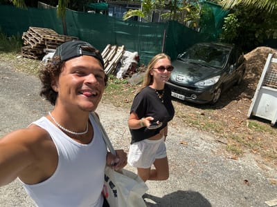 Two people stand on a paved area next to a road with a parked car, likely taking a selfie. The person on the right wears a white tank top and a pearl necklace. The person on the left wears a black top, white shorts and sunglasses, and holds a cell phone. The background includes vegetation, a fence, wooden pallets, and what appears to be a green netting structure. The number plate of the parked car shows the registration "P-323-WA" which identifies the image as being in Sainte-Anne, France.