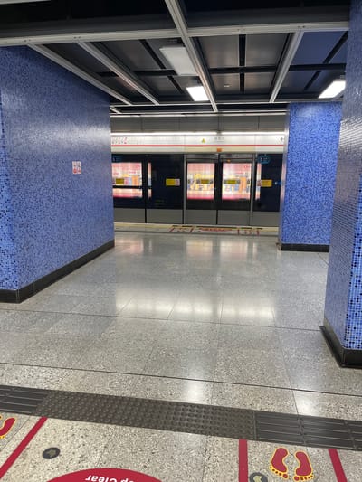 The image shows a view of a subway station platform in Futian District, China. The platform is covered in grey speckled tiles, with tactile paving for the visually impaired running along the edge. The walls of the platform are covered in blue mosaic tiles. On the platform, a subway train is stopped, with its doors open. The train cars have windows and doors with advertisements visible inside. Signs with Chinese characters and pictograms are posted on the walls and doors. There are red and yellow footprints on the floor, indicating a waiting line. The ceiling of the station is dark with a grid of fluorescent lights. The scene appears to be empty of people.