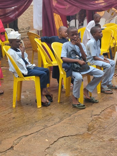 The image captures a group of young males, likely boys or teenagers of African descent, seated on yellow plastic chairs in what appears to be an outdoor or semi-outdoor setting in Unguwar Rimi, Nigeria. The image is oriented vertically.
The subjects are dressed in light-colored, long-sleeved shirts and trousers, mostly in shades of light blue or grey, with one boy wearing dark blue trousers. Some are barefoot or wearing sandals/slides; one black sandal prominently displays a white "B" logo. One boy wears a white embroidered cap (kufi), and another has a black shoulder bag resting on his lap, over which a dark blue cap with partially visible white text, possibly "STRIENT" or similar, is placed.
The scene suggests a gathering or event, indicated by the arrangement of chairs and decorative fabric drapes in maroon/purple, yellow, and white visible on the left side of the frame. The boys are mostly looking forward or slightly upwards, appearing attentive or observing something out of frame. The ground beneath them is a worn, textured concrete or paved surface, showing cracks and what might be dampness or stains.
The bright, natural lighting indicates it is daytime. The overall atmosphere is calm, with no strong emotions discernible.