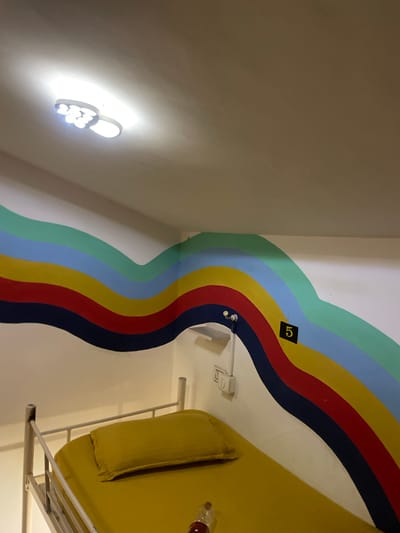 The image shows a close-up interior view of what appears to be a bunk bed in a hostel room. The bunk bed frame is made of metal with a white mattress and a yellow pillow. The wall behind the bed features a vibrant, wavy mural with multiple horizontal stripes of different colors, including navy blue, red, yellow, light blue, and mint green. A small black square with the number "5" in yellow is affixed to the wall, likely indicating a room or bed number. A light fixture with multiple bulbs is visible on the ceiling, and a light switch is mounted on the wall near the bed. The scene suggests a casual, possibly budget-friendly accommodation in Vagator, India, given the context.