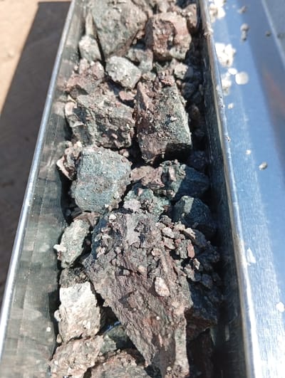 The image shows a close-up, overhead view of mineral ore samples contained within a long, narrow, metallic trough. The trough appears to be made of polished metal, likely stainless steel, and is positioned outdoors under bright sunlight, casting a shadow on the ground to the left. The ore is composed of numerous irregular-shaped rocks and fragments, varying in size from small pebbles to larger chunks. The coloration of the ore is predominantly dark gray and brown, with some pieces exhibiting a distinct greenish or bluish hue, possibly indicating the presence of copper minerals. The texture of the rocks appears rough and granular. The trough is filled with these ore samples, obscuring much of its internal surface. In the background, the ground is visible, appearing to be sandy or dusty. The image is consistent with a geological sampling or assaying process, potentially at the Chelopech mine in Chelopech, Bulgaria, which is known for its copper and gold production. There are no people or visible signs of activity other than the static display of the ore samples.