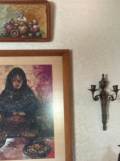 The image shows a portion of a wall in a location likely in Ciudad Victoria, Mexico, based on stylistic cues in the art. The wall is painted with a textured, off-white paint. A metal, antique-style sconce is mounted near the top of the frame. Below the sconce, two framed artworks are partially visible. On the right is a larger, horizontally oriented framed painting featuring dark, abstract figures with splashes of purple and red in the background. To the left is a smaller, vertically oriented painting of a woman carrying pots or vases on her head. Both paintings are within wooden frames of similar color and style. The environment appears to be an interior setting, possibly a living room or gallery.