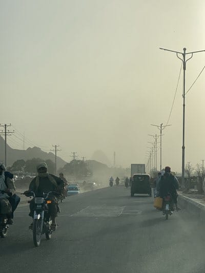 This image captures a bustling multi-lane road scene in Kandahar, Afghanistan, under a heavily hazy or dusty sky. The primary activity is vehicle movement, with a significant number of people commuting or traveling along the road.
The subjects predominantly consist of individuals riding motorcycles, with several motorcycles visible in the foreground and stretching into the distance. One man on a motorcycle in the left foreground wears a face covering, likely for protection against dust. Another motorcyclist in the mid-ground on the right carries a yellow jerrycan on the back of his bike. Beyond the motorcycles, various cars, including a light-colored sedan and a dark SUV/van, and a large commercial truck are also present on the road.
The setting is a wide, paved road flanked by power lines and modern streetlights on both sides. In the background, rugged mountains rise, their forms softened and largely obscured by the thick atmospheric haze, which dominates the upper portion of the image.
Notable details include the pervasive dust or haze, which gives the scene a muted, desaturated quality and significantly reduces visibility into the distance. This environmental condition suggests either a dust storm or typical dry, arid weather. The lighting, soft and diffused, indicates either early morning or late afternoon. The visible infrastructure and types of vehicles, combined with the rugged, dusty mountainous backdrop, align with the provided location context of Kandahar, Afghanistan. No discernible text is visible on any objects or signs within the image.