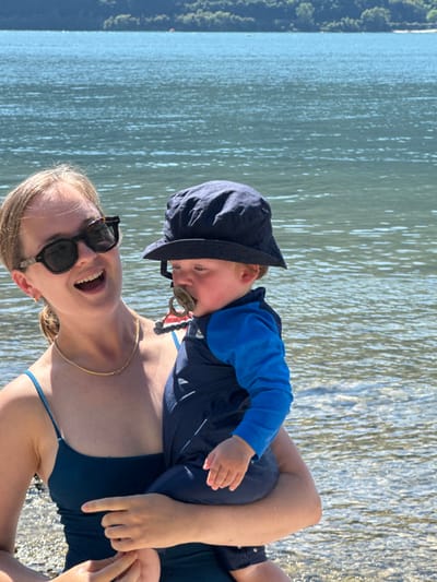 The image depicts a young adult female holding an infant against the backdrop of Lake Como in Abbadia Lariana, Italy. The female, positioned on the left, has light-colored hair, wears tortoise-shell sunglasses, and a dark-colored swimsuit or top with thin straps. She exhibits a wide smile, revealing her teeth, and wears a thin gold chain necklace. Her arms support the infant, who is dressed in a navy blue bucket hat and a two-toned blue and navy long-sleeved rash guard. The infant, with light-colored hair visible, has a brown and red pacifier in its mouth. The background consists of rippled, blue-green water extending to distant, tree-covered hills or mountains under bright daylight conditions.