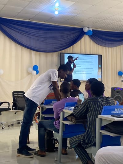 An indoor event is captured in Baganakwo, Nigeria, within a room featuring light-colored walls adorned with horizontal blue and white fabric swags and clusters of blue and white balloons. A large flat-screen display is mounted on the far wall, showing a text-based presentation. A bearded male, wearing a dark t-shirt and a cap, stands in front of the screen, holding a microphone and gesturing towards the display with his right hand.
In the mid-ground, a tall male in a white t-shirt with a partial blue logo and dark jeans leans forward, engaging with a seated male in a purple t-shirt and dark jeans, who occupies a blue and white student desk. Another male participant, dressed in a dark long-sleeved shirt with vertical light stripes and matching pants, is seated at a similar desk, looking down at a smartphone he holds in both hands. Multiple other individuals are partially visible, seated in rows at blue and white desks, generally facing the direction of the presenter. A black portable speaker unit is situated on the floor to the right. The ceiling is white and tiled, featuring a single exposed light fixture.