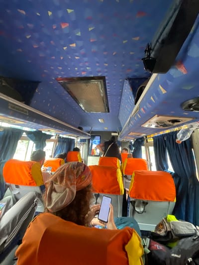 The image depicts the interior of a bus, viewed from a low angle, looking towards the front. The ceiling of the bus is a vibrant blue with a repeating pattern of small, abstract triangular shapes in yellow, red, and white. Along the sides, overhead compartments are visible, lined with blue fabric. Dark blue curtains hang by the windows. Numerous bright orange seat covers with yellow trim are prominent, suggesting a public transportation or tour bus. Several passengers are visible, mostly seen from behind or the side. One passenger in the foreground wears a patterned headscarf and is looking at a smartphone. Another passenger to their left is wearing grey pants with white stripes. Luggage is piled near the seats. The environment appears to be dimly lit, with natural light filtering through the windows. There is no indication of the specific location within Addakal, India, beyond it being the interior of a bus.