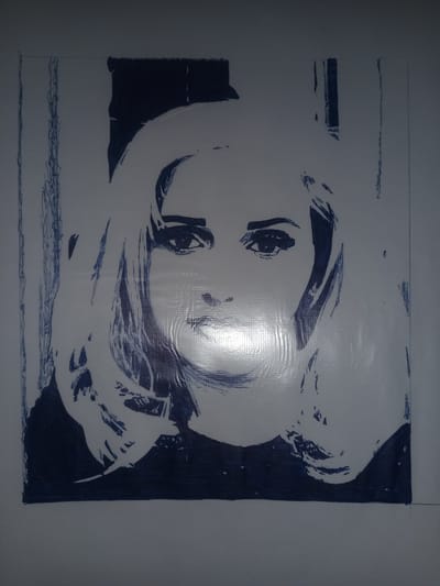 This media file is a photograph of a pen-and-ink drawing on a light-colored surface, possibly paper or a canvas. The drawing is a stylized portrait of a woman with blonde hair, rendered in dark blue ink. The style is reminiscent of pop art, with bold outlines and minimal shading, creating a graphic, high-contrast effect.
The subject is a head-and-shoulders portrait of a woman looking directly at the viewer. Her hair is voluminous and styled in waves. The facial features, particularly the eyes, are emphasized. There is a significant highlight on the bridge of her nose and forehead, suggesting a light source or a reflective quality of the drawing surface. The background is largely white, with some faint vertical lines drawn in blue ink on the left and right sides of the portrait, possibly suggesting a frame or background elements. The bottom of the drawing features a dark, solid area of ink, representing her clothing or a base.
The drawing appears to be relatively recent and is not a historical artifact. There is no visible text within the image. The lighting in the photograph suggests it was taken indoors, possibly in a well-lit room, with the flash of the camera creating the prominent highlight on the drawing. The location context provided is Gulbag, Uzbekistan, which does not have any direct visual cues within the image itself.