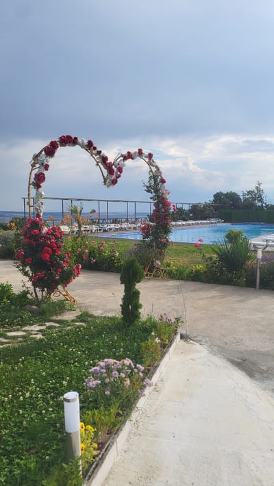 This media file captures a heart-shaped floral arch decorated with red and white roses, positioned outdoors next to a swimming pool. The arch is a prominent object in the scene, with its floral embellishments suggesting a festive or romantic occasion, possibly a wedding or celebration. The setting is a poolside area with paved walkways and green grass. Beyond the arch and pool, there are rows of empty lounge chairs, hinting at a place of leisure. The background reveals a body of water, likely a lake or the sea, with distant trees and hills, under a bright sky with scattered clouds. The time of day appears to be daytime, with ample natural light. The weather seems pleasant, suitable for outdoor activities. There are no people visible in the image. The overall atmosphere is serene and picturesque, emphasizing the beauty of the floral arch and its surrounding environment. The location is identified as Topolitsa, Bulgaria. No visible text is present in the image.