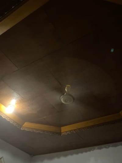 The image shows a ceiling with a dark brown surface, likely wood panels. In the center of the ceiling, a white ceiling fan is visible, with its blades blurred due to motion. To the left, a bright light bulb is illuminated. The ceiling has decorative wooden trim along the edges. There are no people or animals in the image. The scene appears to be indoors, in a room with a ceiling fan and light fixture. The location of Buleleng, Indonesia is not directly depicted or inferable from the image itself, but may be context provided by the user.