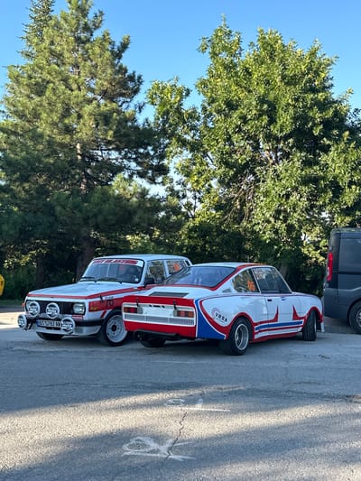 Two vintage rally cars are parked on a paved surface in front of a line of trees under a clear blue sky. The car on the left is a white sedan with red racing stripes and decals, including "HELLA" and "NARVA" branding on its front. It has four auxiliary driving lights mounted on the front bumper. The car on the right is a white coupe with a red and blue racing livery, featuring the number "05" prominently on its side and a "VRBA MOTORSPORT" logo. A dark gray van is parked to the right of the coupe. The ground shows some cracks and chalk markings. The trees are lush and green, suggesting a temperate climate. The overall scene appears to be a parking area, possibly at an event or a private residence, in Lyaskovets, Bulgaria, given the license plate format "BT 5787 AV".