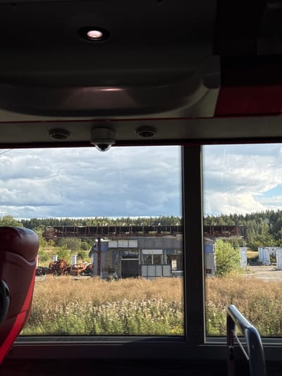 The image depicts a static scene viewed from inside a vehicle, likely a bus or train, looking out a window. The upper third of the frame shows the vehicle's dark ceiling with an illuminated circular light fixture, a central dome-shaped security camera, and two recessed air vents. Below these, the vehicle's interior structure transitions to a window frame. A portion of a red or maroon upholstered seat is visible on the far left, and a silver metal handrail extends from the bottom right corner towards the center.
Through the window, an overgrown, unkempt landscape unfolds under a partly cloudy sky dominated by large cumulus clouds. In the midground, a field of tall, dry, light brown grass and some green vegetation occupies the lower half of the outdoor scene. Beyond this field, several dilapidated structures are visible, consistent with an abandoned industrial or commercial site in Mriia, Ukraine. A prominent, single-story building, possibly a former service station or office, features shattered windows, exposed interior elements, and damaged, discolored walls with remnants of blue and grey paint. To the left of this building, a pile of heavily rusted and mangled vehicle frames, primarily cars, sits among the tall grass. Behind these ground-level structures, an elevated, horizontal metal framework with a partially collapsed roof, resembling a loading platform or conveyor system, spans the scene. Further to the right, several weathered concrete posts or remnants of structures are visible. The background features a dense line of pine trees forming a horizon. No people are present, and no activities or interactions are observed.