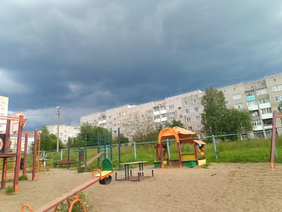 The image depicts a somewhat gloomy outdoor scene, likely a playground in Chusovoy, Russia. The sky is filled with heavy, dark clouds, suggesting an impending storm or recent rainfall.

In the foreground, a sandy playground is visible, featuring various play structures. There's a seesaw with orange handles and a green and yellow seat. To the right, a bright orange and red sandbox shelter with a yellow roof stands, housing a small, colorful play structure within. A picnic table with benches is also present. Further back, more playground equipment, including swings and climbing frames, can be seen. A red wooden bench is situated near a metal fence.

Behind the playground, a line of tall, multi-story residential buildings with light-colored brick facades and numerous windows stretches across the middle ground. The buildings appear to be typical of Soviet-era apartment blocks. Trees and green grass border the playground and the buildings. A single lamppost stands prominently on the right side of the image, near the buildings.

The overall mood is somber, amplified by the dark clouds and the absence of people. The scene captures a quiet moment in a residential area, likely during a dull or inclement weather day. The visible text on the sandbox shelter reads "KVINTA".