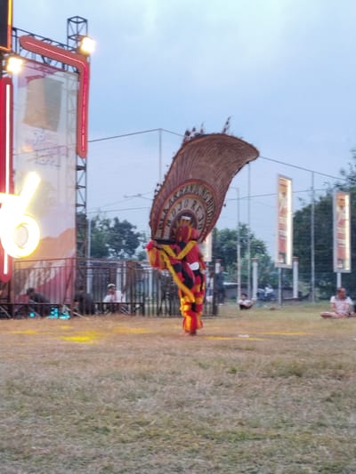 The image depicts an outdoor event, possibly a festival or performance, taking place in Sambirejo, Indonesia. The central focus is a performer in a traditional costume, characterized by a large, fan-like headdress made of woven material. The costume itself is vibrant, with red and yellow accents. The performer is in motion, suggesting a dance or theatrical presentation.

In the background, a large stage structure is visible, adorned with bright lights, including neon-red and yellow elements. A prominent, illuminated "6" is part of the stage design. Behind the stage, a chain-link fence encloses an area, and beyond that, trees and a cloudy sky are present. Several vertical banners with illuminated borders are also visible in the middle ground, some displaying text.

On the grassy field in front of the stage, a few spectators are seated or kneeling. Some are observing the performance, while others appear to be resting. The ground is covered in dry grass, with scattered patches of yellow powder or material. The overall atmosphere suggests a lively cultural gathering.