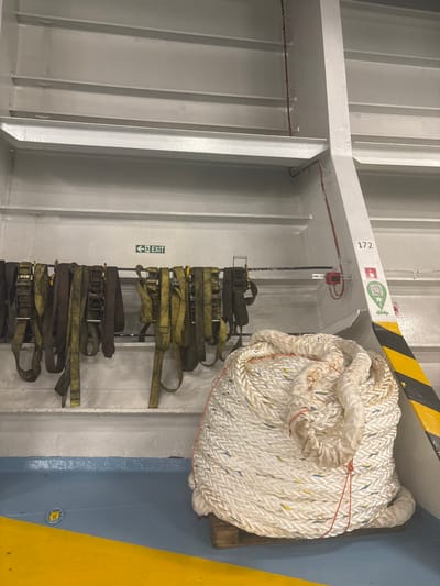 A white rope coil sits secured to a painted blue wall with yellow trim. Below it, on a metal rack attached to a white wall with vertical beams, are multiple stacked, dark yellow cargo straps. An "EXIT" sign is visible. The wall above the rope has a red sensor and a green and red sticker marked "172." A yellow and black hazard stripe is at the top.