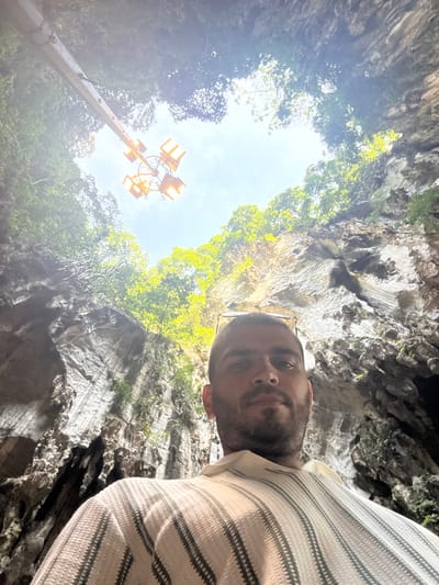 The image presents an upward-looking perspective from within a large natural rock formation, likely a cave or a deep sinkhole, under a bright sky. In the foreground, partially visible on the right, is a man looking towards the camera. He has a short beard and wears glasses perched on his head. He is dressed in a light-colored, possibly off-white, top with horizontal dark stripes.

The scene is dominated by the rugged, grey-brown rock walls of the cave, displaying natural textures and crevices. Green foliage and trees are visible, growing from the rock surfaces and outlining the circular opening at the top, through which a clear, bright blue sky is seen. A tall metal pole with several bright yellow-orange floodlights extends upwards from the lower-left, providing artificial illumination to the natural environment.

The activity depicted is a personal photograph, possibly a selfie, taken by the man or someone directly below him, during a visit to this natural site. The bright sky suggests it is daytime, likely late morning or early afternoon. The location, given the context of Selayang Municipal Council, Malaysia, indicates this is a natural attraction within that area, possibly a well-maintained tourist spot due to the presence of the floodlights. There is no visible text in the image.