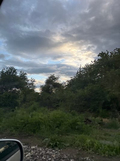 The image captures a view from a vehicle in what appears to be a rural location near Baylovo, Bulgaria. The sky is heavily overcast with a mix of dark and light clouds, with a patch of light filtering through. Trees line the right side of the frame, with dense green foliage. A dirt or gravel road runs alongside the trees, transitioning to a rocky area. The side mirror of the vehicle is visible in the bottom right corner.