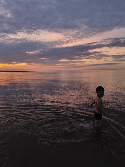 A young boy, appearing to be around six years old, stands in the shallow waters of a large body of water, likely the Baltic Sea, at dusk. He is wearing dark swim trunks and is facing away from the camera, looking back over his shoulder with a slight smile. The water is calm with small ripples, reflecting the colorful sky above. The sky is a blend of deep purples, pinks, and oranges, with dramatic cloud formations suggesting the sun has recently set or is about to rise. The horizon line is clearly visible, separating the water from a faint, dark landmass in the distance. The overall scene is serene and evokes a sense of peaceful solitude at the beach. The location is identified as Apšuciems, Latvia.