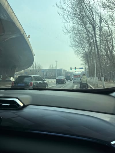 The image displays an urban road scene viewed from the interior of a vehicle, looking forward through the windshield. The setting is Huanggu District, China, under an overcast or hazy sky during daylight hours.

In the immediate foreground, the dark gray dashboard of the viewer's vehicle is visible, featuring a subtly patterned surface and a chrome-accented air vent on the left.

The road ahead is a multi-lane thoroughfare with vehicular traffic. A significant concrete overpass structure curves across the upper left portion of the frame. To the right, a line of leafless deciduous trees borders the road. Traffic lights ahead are illuminated green, indicating an open intersection.

In the midground, directly in the lane ahead, a person wearing a light-colored (beige or cream) puffy jacket is operating a small, light-colored scooter or motorcycle. Immediately beyond the scooter, a white van with red Chinese characters on its rear is visible. In the adjacent left lane, a dark green four-door Mini Cooper, bearing the Chinese license plate "辽A 102OM," is positioned. Multiple other dark-colored sedans and vehicles are present in various lanes further down the road. White road markings, including an arrow indicating straight movement, are discernible on the asphalt. Industrial or commercial buildings are visible in the distant background.
