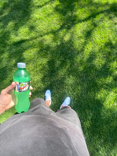 A person stands on a lawn wearing blue shoes and gray pants. They hold a green bottle of Squirt soda. The lawn is green and appears to be partially shaded. The view is from above, looking down at the person and the ground.
