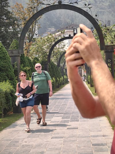 The image captures a paved pathway in Sa Pa, Vietnam, extending into the background beneath a series of dark, ornate metal arches. In the immediate foreground, the arms and hands of a person hold a mobile phone horizontally, pointing it towards the midground subjects.
Two individuals are walking on the pathway towards the viewer. The female on the left wears a black tank top, denim shorts, and light-colored flat sandals. She has sunglasses on, a dark backpack, and a white fanny pack, and carries a clear plastic water bottle along with a white patterned item. The male on the right wears a green "Timberland" t-shirt, dark shorts, and dark brown two-strap sandals. He also wears sunglasses and a dark backpack. Both individuals are walking side-by-side.
The pathway is composed of rectangular grey paving stones. On either side, manicured green bushes and grass strips flank the path, leading up to the decorative metal arches. Beyond the arches, a landscape of green trees and hazy hills is visible, interspersed with buildings, one of which exhibits a tiered roof design typical of traditional Asian architecture. The scene is illuminated by direct sunlight, casting shadows on the ground.