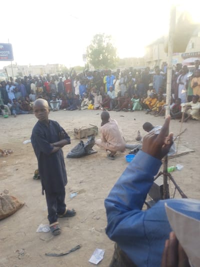 An outdoor scene in Babban Giji, Nigeria, depicts a large informal gathering of individuals on a dusty, unpaved ground. In the foreground, slightly left of center, a young boy with short hair, wearing a dark blue traditional garment, stands facing the camera with his arms crossed. To his right, in the mid-ground, a man in a light brown traditional outfit crouches, facing away from the camera, attending to items on the ground, including a wooden crate, a black plastic bag, and a blue plastic bowl. In the extreme lower right foreground, the arm and shoulder of an individual in a blue long-sleeved shirt and a white cap are visible, with their right index finger raised vertically.

The background features a dense crowd of approximately 50-100 people, predominantly men and boys, many dressed in traditional Nigerian attire, seated and standing in a semi-circular formation, observing an unseen central point. To the far left, a blue sign with white lettering displaying "DANGOTE CEMENT" is visible. Behind the crowd, low-rise buildings and a single tree are discernible under bright daylight conditions. The ground is littered with miscellaneous debris, including plastic bottles, paper, and small plastic bags.