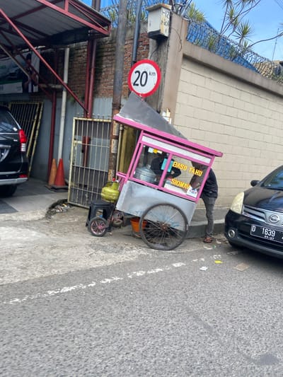 The image shows a street food vendor selling "Baso Tahu Siomay" in Kecamatan Margahayu, Indonesia, located at coordinates -6.963 latitude and 107.578 longitude. The vendor is a man standing next to a pink and grey food cart. The cart has a large wheel on the right side and a smaller wheel on the left side. There is a black car parked in front of the vendor. The car has the license plate "D 1639." The vendor's cart is positioned next to a brick wall with a sign that says "20 km."  There is a yellow propane tank connected to the cart. There is also a small stool next to the cart. The vendor is wearing a black shirt and black pants. The street appears to be paved and has a white line down the center. The sky is blue and there are trees in the background.