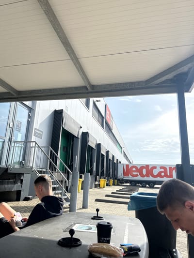 The image shows a low-angle view of a large commercial building, likely a distribution center, located in Ede, Netherlands. The building has multiple loading docks with black canopies extending over them. A "Nedcargo" semi-truck is parked in front of one of the docks. A metal staircase provides access to one of the loading docks. A sign reading "EasyWork-" is visible near a window.
Two people are partially visible in the foreground. They appear to be sitting at a table with coffee cups and food. They seem to be at an outdoor break area or patio. The environment includes a clear sky.
