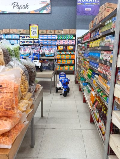 The image captures an eye-level view down a well-lit aisle of a supermarket or grocery store. No people are visible, and the scene is still.
On the left side of the foreground, a display table is laden with numerous clear plastic bags filled with various types of packaged snacks, such as puffed corn snacks and crackers. Beyond this, another table holds more items, including children's ride-on toys – one resembling a white and grey car, and another blue and white tricycle-like toy with "POLICE" written on it. This blue and white toy is positioned centrally in the aisle, facing away from the camera.
The right side of the aisle features tall metal shelving units stocked with an array of bottled beverages, including water, juices, and various flavored drinks, arranged neatly in rows. The items on these shelves extend deep into the background.
At the back of the aisle, large shelves are filled with packaged goods, predominantly diapers and baby products, with brands like "Nice" visible on packs of tissue paper. Cardboard boxes are stacked on top of these shelves. A yellow price sign prominently displays "249.000,-".
Above the back shelves, a large white banner with "Popok" (Indonesian for 'diapers') and "Robinson" branding is visible. Another purple banner further back reads "MAHAL, UANG KEMBALI!!" (Expensive, Money Back!!) also with the "Robinson" logo, indicating a promotional offer within a Robinson store, common in Indonesia given the language and currency context. The floor consists of light-colored tiles. The scene suggests a retail environment focusing on baby care, snacks, and beverages.