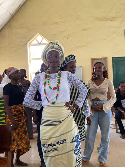 This media file depicts a group of young women in what appears to be an indoor setting, possibly a classroom or community hall in Dutsen Kato, Nigeria. The image is oriented vertically, showing several individuals from the chest down.
The primary subject is a young woman in the foreground, wearing a white lace top and a headscarf. She is adorned with a colorful beaded necklace featuring red, yellow, green, and black beads. She is also wearing a fabric draped around her, with black and white stripes and visible text that appears to be in a non-Latin script. Beside her, other women are visible, some wearing headscarves and patterned clothing. One woman in the background wears a light-colored top with a butterfly design and blue jeans. The scene seems to be a gathering or an event, possibly a performance or a cultural presentation, given the attire and the focus on the foreground subject.
The setting is characterized by a textured, light-colored wall, a small window with a wooden frame, and a tiled floor. The lighting suggests it is daytime, with natural light coming from the window. The overall mood appears to be one of communal gathering and perhaps pride or celebration, as indicated by the celebratory attire of the women. There is no discernible information about the weather.
Visible text on the fabric in the foreground reads "GBAGIGHI BECHEH" and "AZAR (XORO)". The context of Dutsen Kato, Nigeria, suggests the cultural significance of the attire and the event.