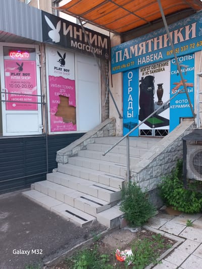 The image shows a storefront in Volzhsky, Russia featuring two adjacent businesses. On the left is an "Intim Magazin" or intimate shop, named "Playboy Intim Shop", with a sign that indicates opening hours of 10:00-20:00, and sells intimate products for adults and lingerie. To the right is a shop selling tombstones and monuments. The sign on the second shop is advertising tombstones, monument installation, table legs and ceramic photos. In front of the stores are stairs and sidewalk. Some greenery is growing in the sidewalk.