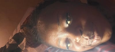 The image shows a close-up of a person's face, lying down. The person appears to be resting on a patterned surface, possibly a pillow or blanket. The lighting in the image is dim, with a warm glow illuminating the person's face. The person's eyes are open and looking towards the side, with long eyelashes visible. The skin appears to have a natural sheen. The hair is braided and pulled back. The image is oriented sideways. There is no discernible information in the image that would allow for the determination of its location in Masaka, Nigeria.