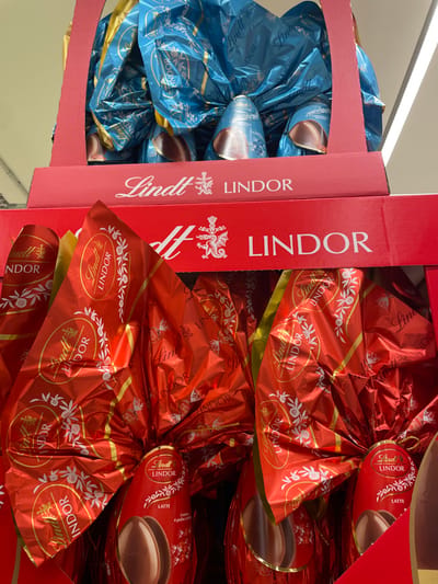 This image presents a close-up view of stacked retail display boxes containing Lindt Lindor chocolate Easter eggs within a commercial establishment in Monsummano Terme, Italy. The lower, foreground display box is red cardboard and holds multiple large chocolate eggs individually wrapped in bright red foil. Each red-wrapped egg features "Lindt LINDOR" branding and decorative white patterns, with the visible product labels indicating "LATTE" (milk chocolate) and showing a depiction of the chocolate egg itself. Above this, another red cardboard display box contains large chocolate eggs wrapped in vibrant blue foil, also bearing "Lindt LINDOR" branding. The blue-wrapped eggs exhibit portions of a lighter blue inner wrapper or the chocolate shell visible at their bases. Both display boxes are clearly labeled "Lindt LINDOR" with an accompanying heraldic crest. The immediate environment is an indoor retail setting, illuminated by overhead lighting, with no individuals visible in the frame.