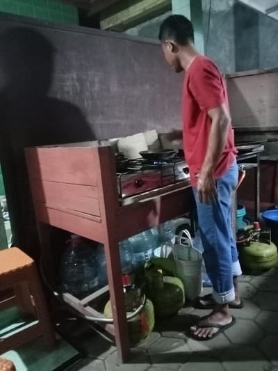 A man wearing a red shirt and blue jeans is standing in a kitchen located in Kecamatan Brangsong, Indonesia, at coordinates -6.943, 110.221. He is standing beside a wooden countertop with a gas stove on it. Under the countertop, there are a few water jugs and green propane tanks. There is a black frying pan on the stove with a small amount of food in it. The man appears to be watching the food cook. The kitchen walls are made of dark brown wood paneling and there is a large black chalkboard on the wall to the left of the man. There is a green tiled wall in the background to the left of the frame and a shadow on the wall behind the man. The floor is made of tile. To the left of the stove is a stool made of woven plastic. The man is holding a small, white object in his hand.