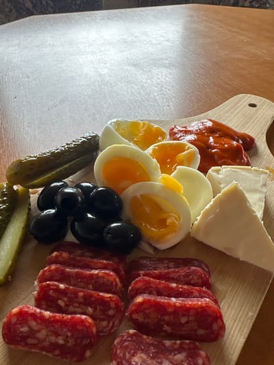 A light-colored wooden cutting board, featuring a handle cutout in the upper right, is depicted resting on a lighter brown wooden table surface in Fowlis Easter, United Kingdom. The board is laden with an assortment of food items. On the left side, two whole gherkins are positioned, one of which is sliced longitudinally to reveal its pale green interior. Adjacent to these, a cluster of approximately ten to twelve whole black olives is visible. Multiple halves of soft-boiled eggs, displaying bright orange yolks and white albumen, occupy the upper-central section of the board. To the upper right, a portion of thick, reddish-orange paste or sauce is present. Below this, two pieces of white cheese, one triangular and one rectangular, are arranged. Along the bottom edge of the board, several short, cylindrical pieces of cured sausage or salami are laid out in rows, featuring varying shades of red and white fat marbling. No individuals, direct actions, or interactions are observable within the scene. The background above the wooden table surface is out of focus, revealing a subtle, textured pattern.
