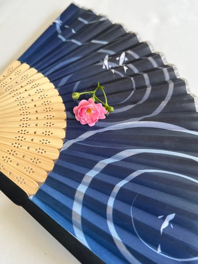 The image displays a close-up, top-down view of an open, traditional Japanese-style folding fan. The fan has a bamboo handle that transitions into a fabric panel adorned with a dark blue background featuring abstract, swirling white lines and subtle, light blue accents. The fabric is pleated, creating a textured surface. Resting on the fan is a single, small, pink rose with a green stem and bud. The background is a plain white surface, suggesting an indoor setting. The scene is static and does not depict any activity or interaction. The location of Kusnatqazma, Azerbaijan is not relevant to the visual content of the image.