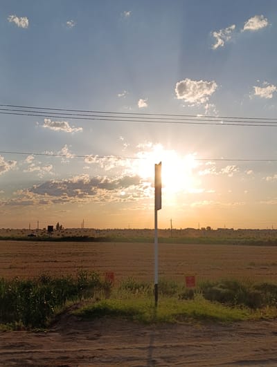 This is a photograph of a rural landscape in the city of Zhañatalap, Kazakhstan, taken during sunset. The sun is bright and low in the sky, casting a warm glow over the scene and creating visible sunbeams. The sky is a blend of blue and orange, with scattered white clouds. In the foreground, there is a dirt path leading towards a field of harvested crops, with some green vegetation along the edge of the path. A white post with a dark top stands in the middle of the frame, in front of the field. In the distance, there are hay bales scattered across the field, and several power lines stretch across the horizon. The overall atmosphere is peaceful and serene, typical of a rural sunset.
