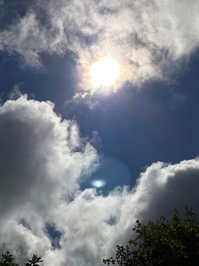 The image is a photograph of a sky with scattered clouds and the sun visible. The sun is bright and appears to be partially obscured by clouds, creating a luminous effect. There is a noticeable lens flare, a circular bright spot, in the lower half of the image. The clouds are varied in texture and density, with some appearing white and fluffy, while others are darker and more diffused. A portion of green foliage, likely from trees, is visible in the lower right corner of the frame, suggesting an outdoor setting. The overall impression is of a dynamic sky on a day with mixed weather. The location is identified as Rathvilly, Ireland.