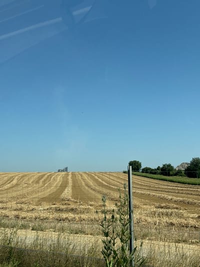 A harvested field occupies the right half of the frame, marked by parallel lines of cut straw. A blue tractor sits in the field. A metal pole and fence run along the field's edge, with some green vegetation growing near them. Trees line the top edge of the field. A clear blue sky fills the left half of the frame, with a faint reflection visible in the bottom left corner.
