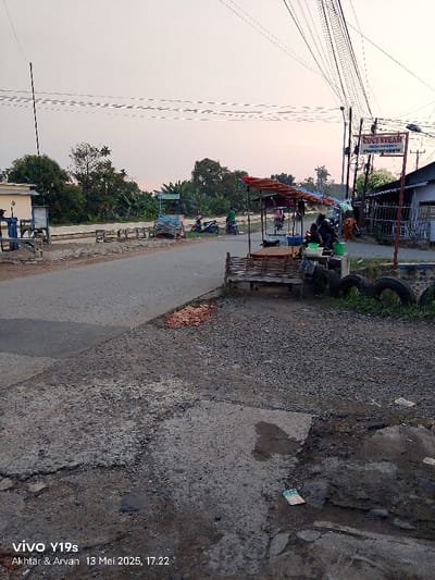 The image shows a street in Kecamatan Kroya, Indonesia, with a small market stand on the right side of the road. The stand has a wooden frame and a blue tarp roof. There is a small wooden bench in front of the stand. There are several people standing near the stand. The road is paved and appears to be in good condition. There are trees and bushes on both sides of the road, with the foliage appearing lush and healthy. The sky is overcast and the image was likely taken in the early evening. The image is taken from the road and points towards the market stand. There are several power lines running along the road. There is a sign that says "DUCI STEAM" in white letters on a blue background, which appears to be a business name. The image was taken with a phone camera, the phone model is a Vivo Y19s. The date and time of the photo are "13 Mei 2025, 17.22".