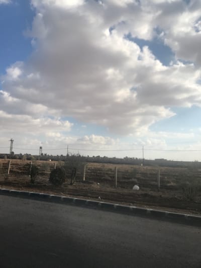 The image depicts an open, semi-developed landscape in Heteitet at-Turkman, Syria, under a partly cloudy sky. In the foreground, a dark asphalt road is partially visible on the bottom left, bordered by a segmented curb of dark and lighter grey blocks. Immediately beyond the curb, a strip of disturbed, dark brown soil with linear patterns is present. This transitions into an uncultivated field characterized by dry, sparse vegetation and exposed light brown earth. Within this field, several small trees or large bushes with dark foliage are visible, along with three light-colored, vertical fence posts. A single white plastic bag lies on the ground in the field. The background features a low, continuous line of indistinct grey structures or walls extending horizontally across the frame. On the left side of the background, a dark metal lattice tower stands beside a light-colored water tank on a support structure. Multiple utility poles with horizontal lines traverse the landscape in the distance. The sky, occupying the upper two-thirds of the image, is filled with numerous white and grey cumulus clouds, with intermittent patches of clear blue sky visible. Natural daylight illuminates the scene. No individuals or active subjects are present.